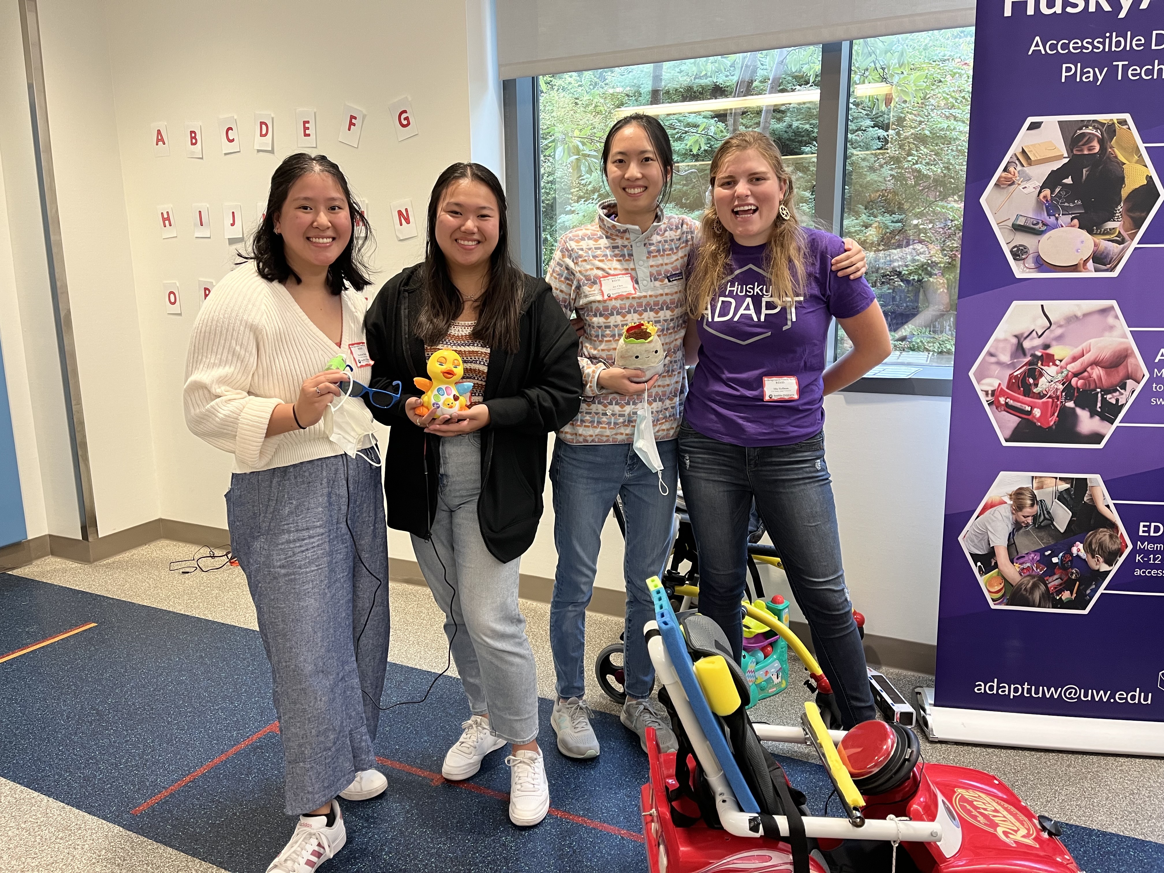 Mia standing with other members of the HuskyADAPT Executive team smiling with switch-adapted toys and an adapted ride-on car.