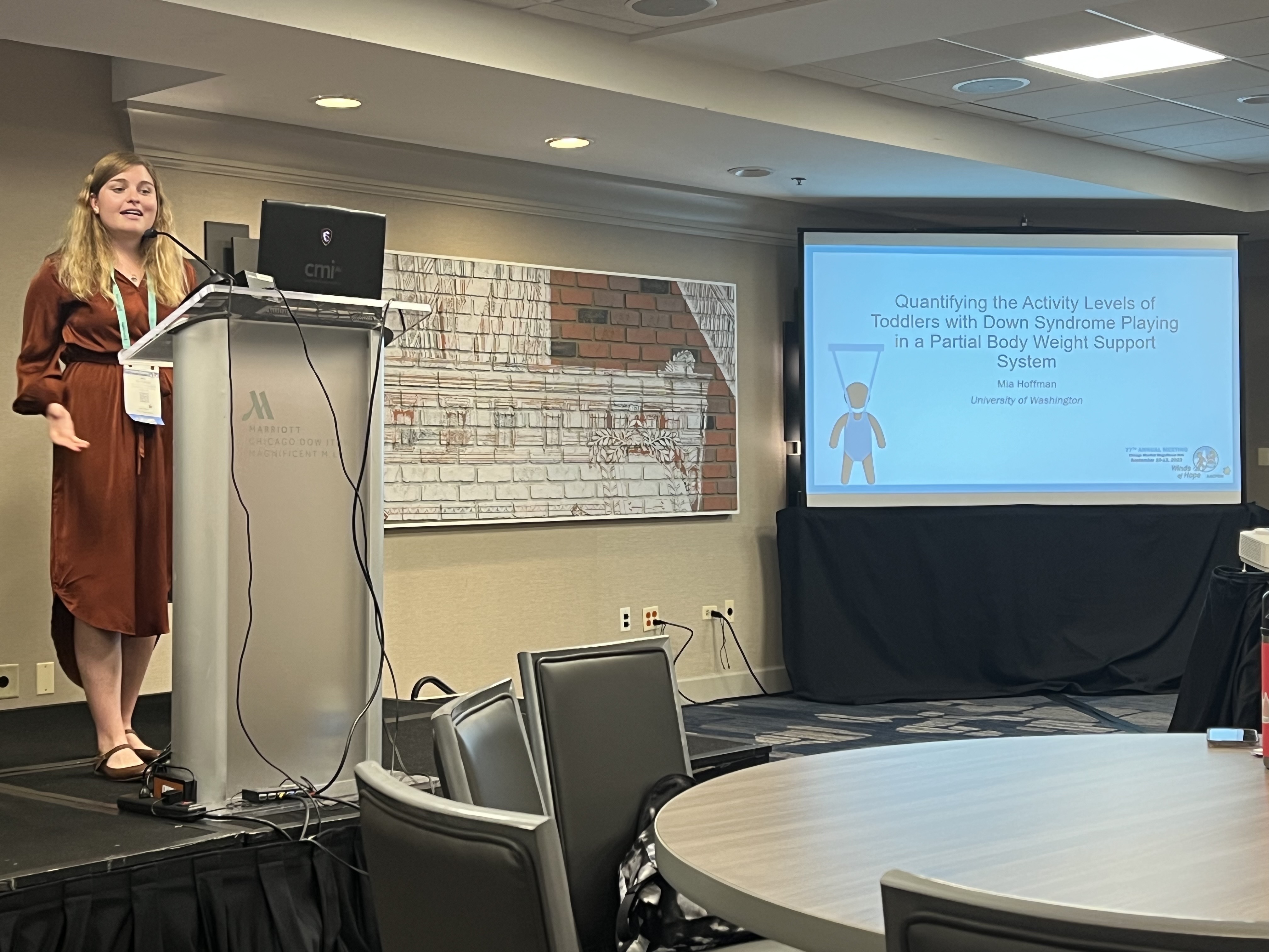 Mia wearing a knee length rust-colored dress presenting a presentation titled Quantifying the Activity levels of toddlers with Down syndrome playing in a partial body weight support system.