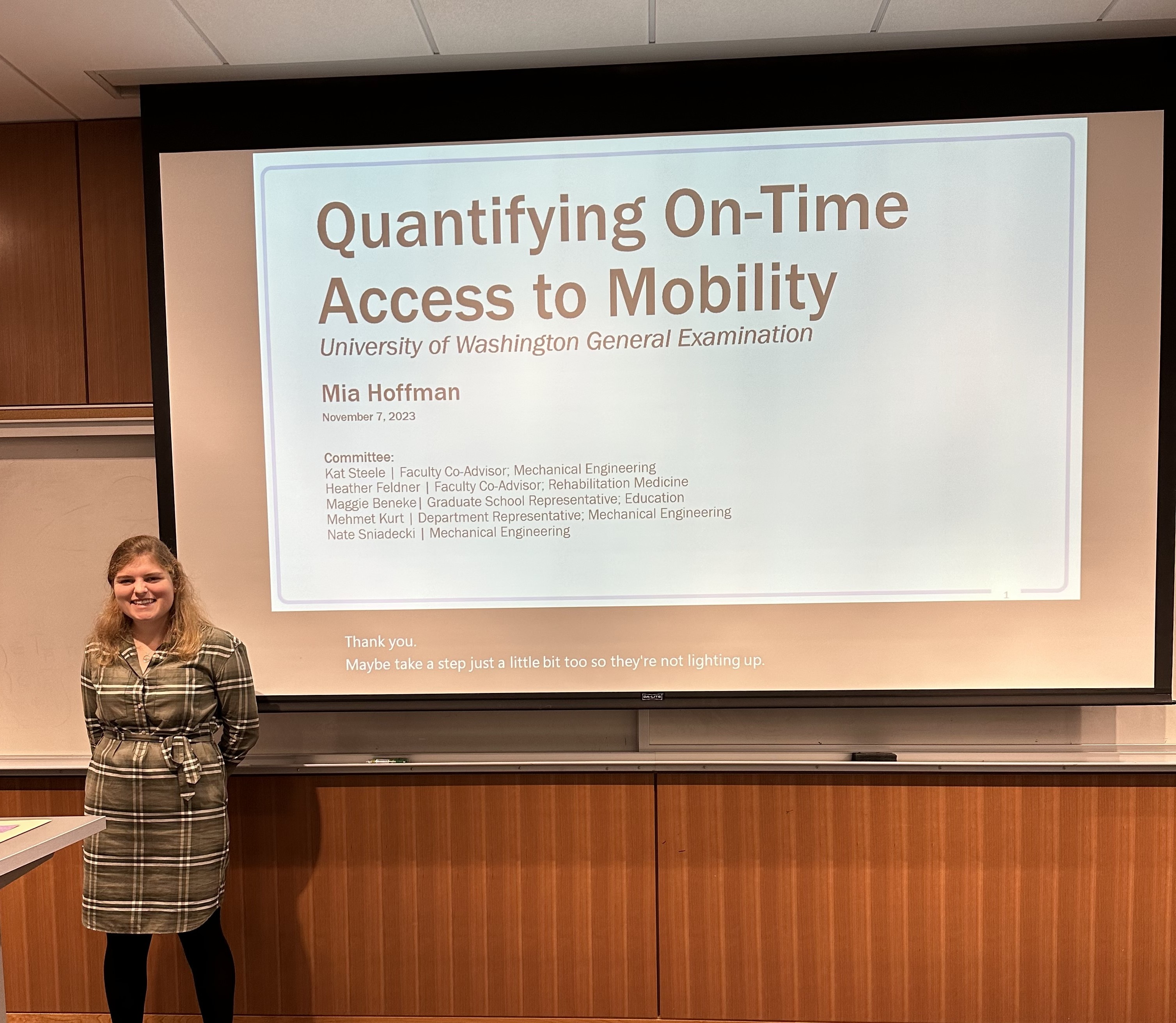 Mia wearing a green plaid dress standing next to a powerpoint reading Quantifying On Time Access to Mobility.