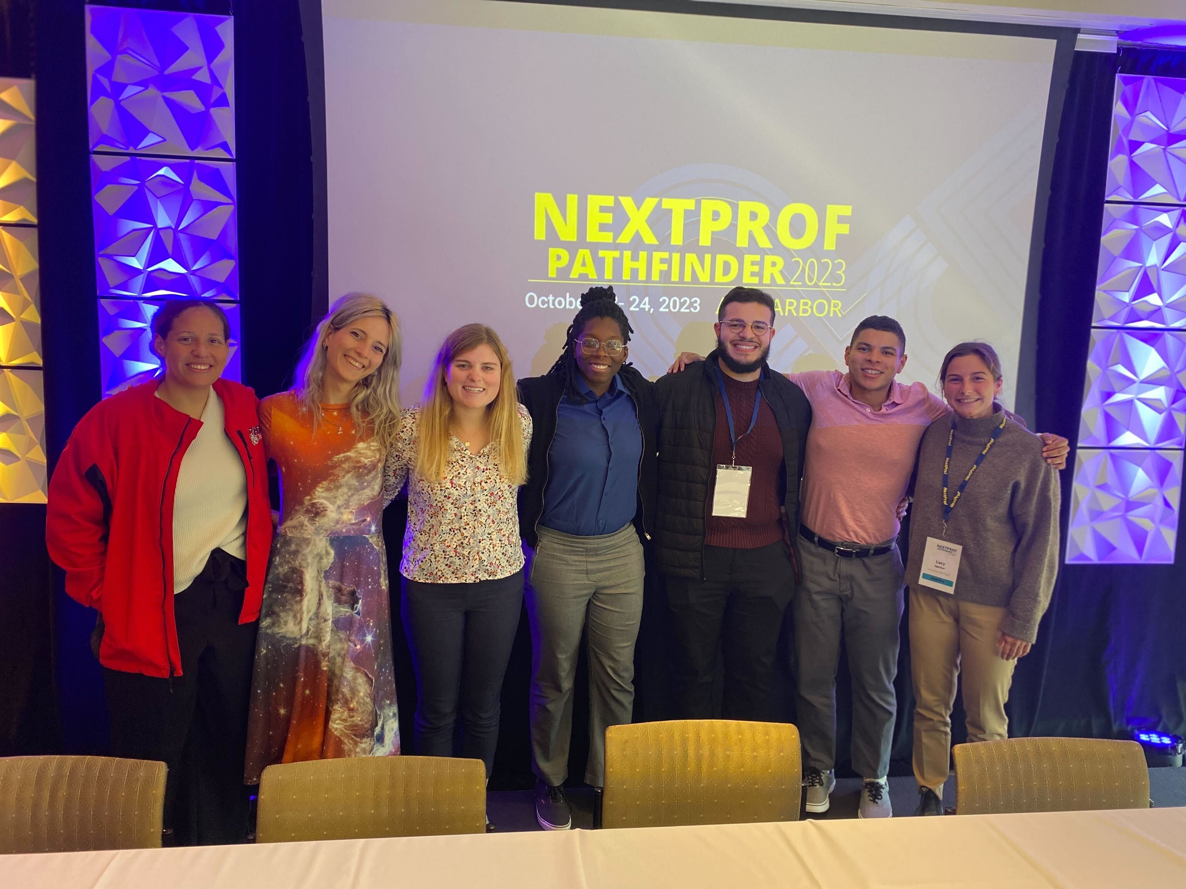 Mia standing with five other PhD students in front a presenting screen that says NextProf Pathfinder.