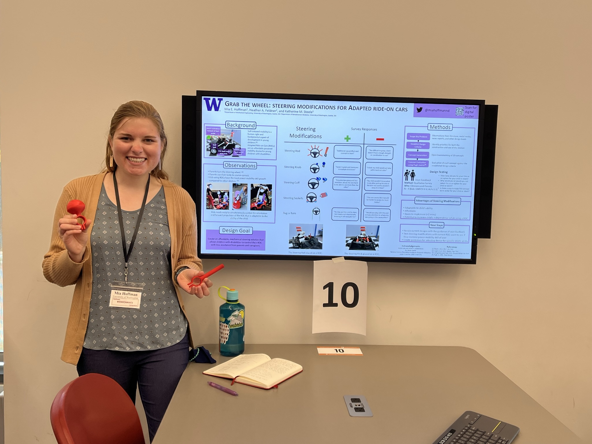 Mia presenting a steering modifications poster on a tv monitor at NWBS while holding 2 3D printed steering modifications.