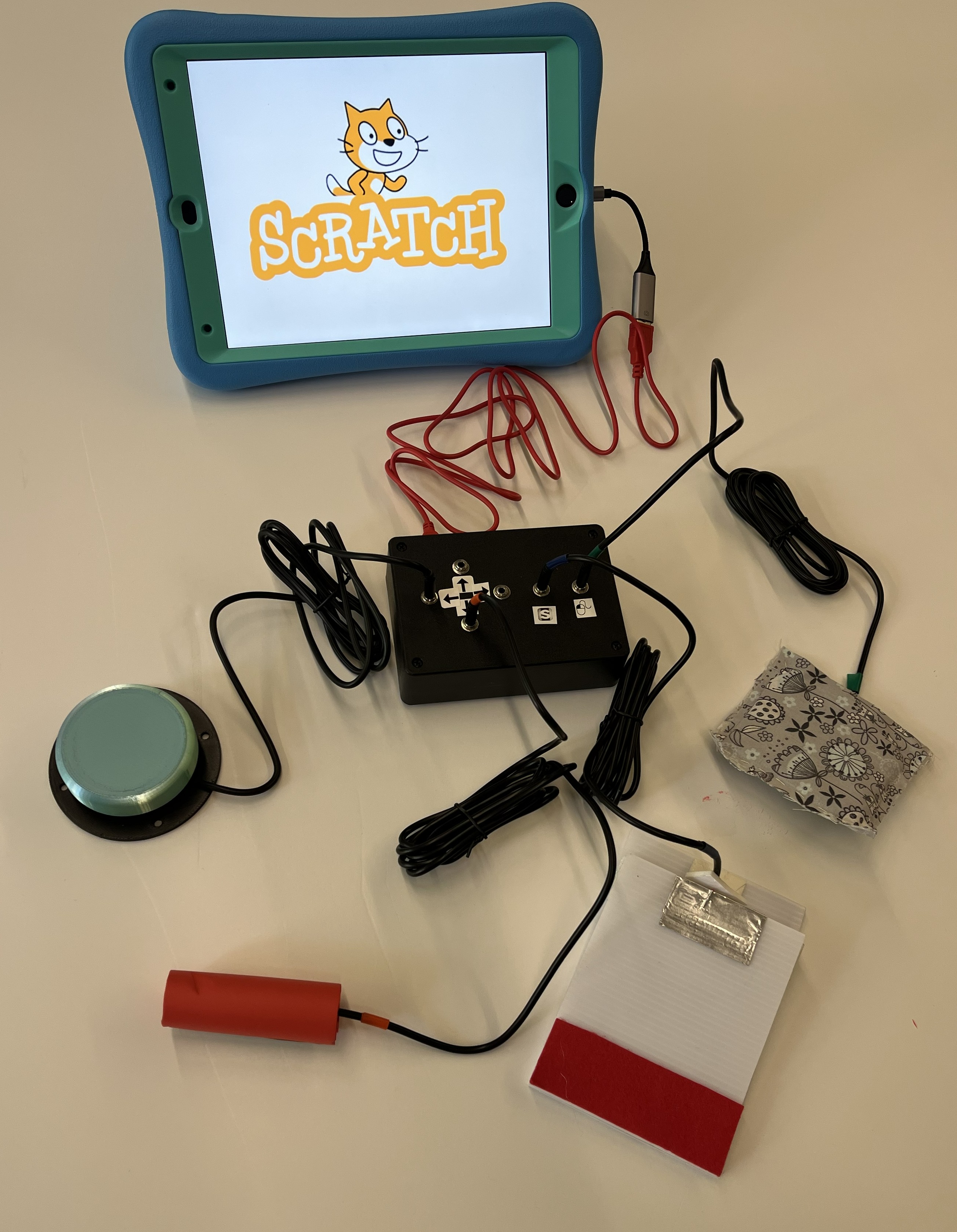The Switch Kit consists of an input device in a black adapted housing, connected to an iPad showing Scratch games, and switches made out of materials like pool noodle plugged into the input device.